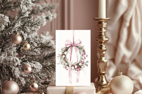 A pastel pink Christmas setup featuring ornaments, a tall candle, and a pink gift box topped with a blank holiday card, with a decorated Christmas tree in the background.