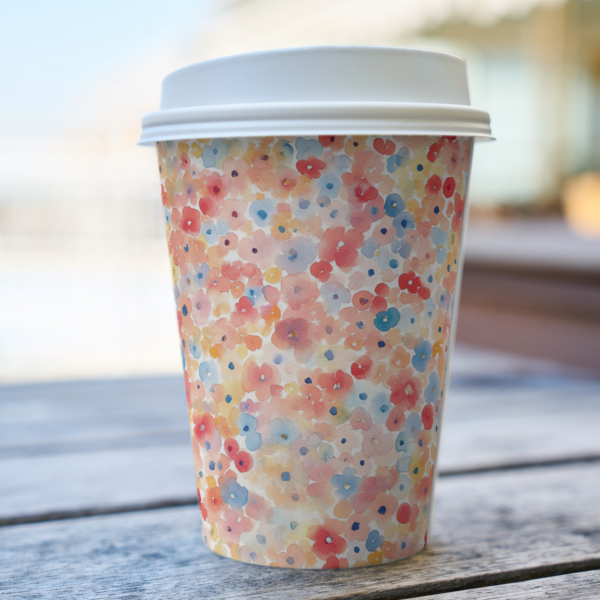 A disposable coffee cup with colorful floral patterns from the Spring Watercolor Seamless Patterns collection is placed on a wooden table outdoors. It is perfect for adding elegance to branding or everyday items.