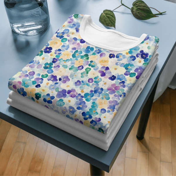 Three folded cotton shirts are on a blue table, and the top shirt features blue, purple, and green floral patterns from the Spring Watercolor Seamless Patterns collection.