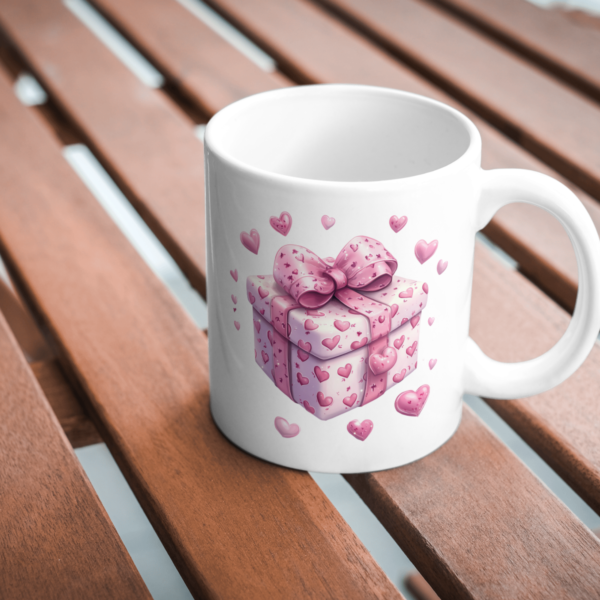 A white mug and pink Valentine’s gift box with heart patterns are displayed on a wooden table, creating a romantic vibe.