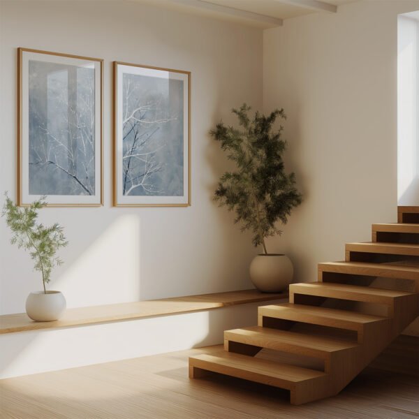 Modern interior with wooden staircase, sunlit white walls, and two framed tree paintings. Green potted plants add contrast and serenity.