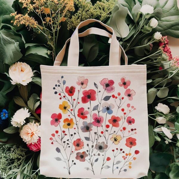 A beige canvas tote bag with colorful painted flowers in red, pink, yellow, and blue, surrounded by real flowers and greenery.