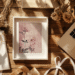 A floral art print rests on a wooden desk surrounded by a laptop, dried flowers, ribbons, and notebooks. Warm sunlight enhances the cozy scene. How to use a digital download from Etsy.