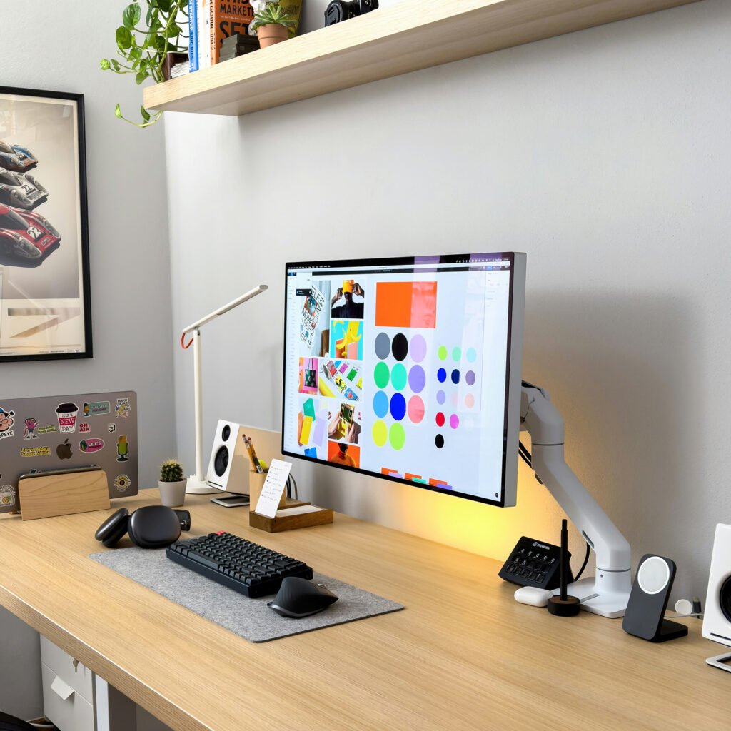 Modern workspace styled with fall moodboard ideas, featuring a large monitor displaying colorful design elements, a tidy desk with speakers, keyboard, mouse, plants, and cozy accessories.