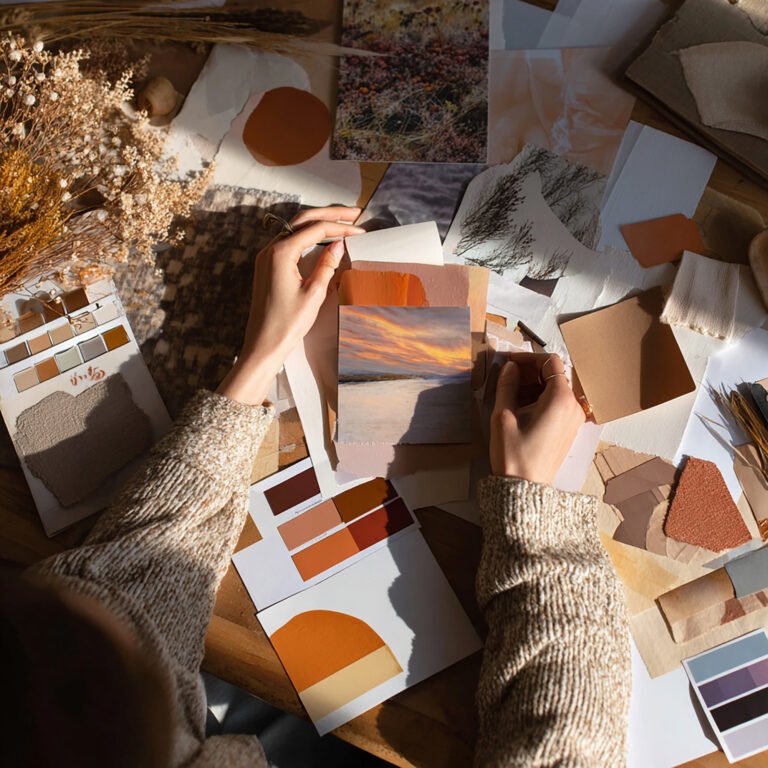 A person arranges paint swatches and photos on a wooden table, styled with fall moodboard ideas in warm earth tones. The flatlay scene conveys creativity, focus, and autumn inspiration.
